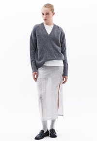 Szary sweter dzianinowy z dekoltem w kształcie V, nałożony na biały top, zestawiony z przezroczystą spódnicą z srebrnymi cekinami z bocznym rozcięciem oraz czarnymi loafersami.