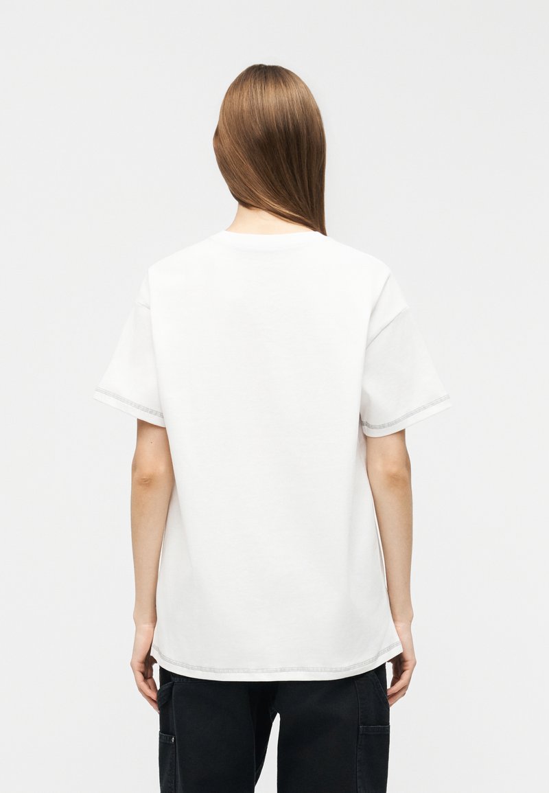 Persona con lunghi capelli castani che indossa una t-shirt bianca a maniche corte semplice e pantaloni neri, di spalle su uno sfondo semplice.