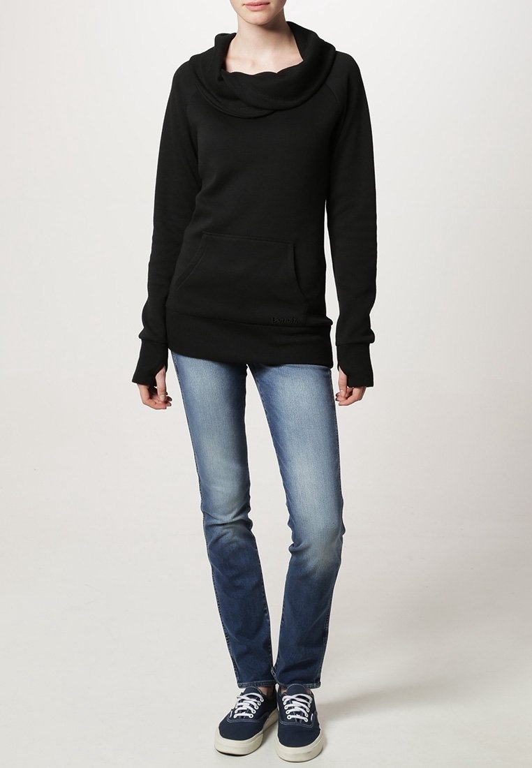 Personne portant un sweat noir à col bénitier avec des trous pour les pouces, un jean bleu délavé et des baskets bleu marine avec des lacets blancs, debout devant un fond uni.