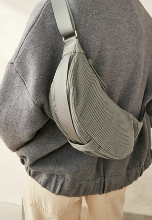 Groene corduroy crossbodytas met een getextureerd oppervlak, verstelbare riem en een afgeronde vorm, gedragen over een grijze trui.