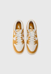 Nike tenisky s bielym koženým zvrškom a žltými semišovými akcentmi, okrúhlym nosom, perforovanými detailmi a bielymi šnúrkami na bielom pozadí.
