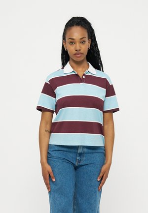 Γυναίκα με μακριά πλεξούδα, ντυμένη με μπλε και μαρόν ριγέ πουκάμισο πόλο και μπλε τζιν, που στέκεται μπροστά σε απλό λευκό φόντο.