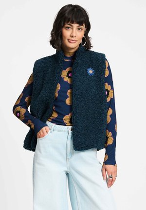 Vrouw met donker haar draagt een blauw bloemenpatroon blouse met lange mouwen, een marineblauw fleecevest met een blauwe broche, lichtblauwe jeans en zilveren oorringen.