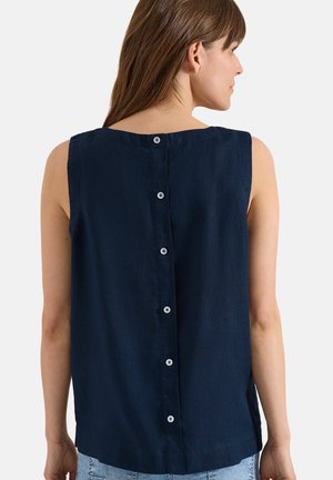 Vrouw draagt een mouwloos donkerblauw topje met witte knopen die verticaal over de rug lopen, gecombineerd met lichtblauwe spijkerbroek.