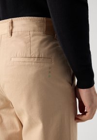 Beige bomulds-chinos med en glat tekstur, prydet med et brunt lædermærke, baglomme og grønne syninger.