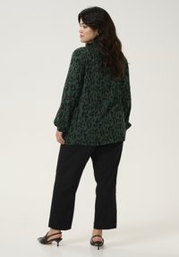 Green patterned blouse with long sleeves, featuring black abstract shapes. Worn with black straight-leg trousers and black heeled mules.
