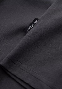 Tmavě šedé bavlněné tričko se hladkým povrchem, s viditelnou černou nášivkou s nápisem "ombre" blízko lemu a s úhledným šitím na okrajích.