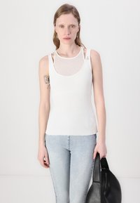 Canotta bianca a coste con sovrapposizione in rete, scollo rotondo, spalline sottili e design aderente, abbinata a jeans azzurri chiari e una borsa nera.