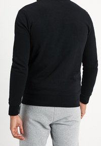 Svart sweatshirt sedd bakifrån, med långa ärmar, ribbade muddar och en enkel design. I kombination med grå sweatpants.