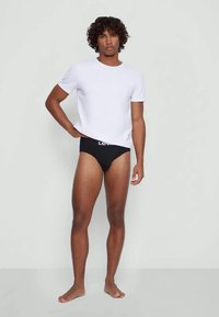 Fehér pamut póló, fekete alsónadrággal, amelynek texturált derékrésze van. A modell mezítláb, egyenes lábakkal áll egy semleges háttér előtt.