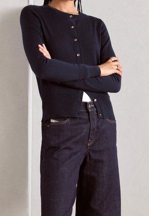 Persona con le braccia incrociate che indossa un cardigan blu scuro abbottonato e jeans in denim scuro su uno sfondo chiaro uniforme.