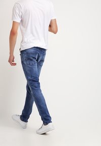 Jeans de mezclilla azul con un corte ajustado, que cuentan con dos bolsillos traseros y un ligero desgaste. Combinados con una camiseta blanca lisa y zapatillas blancas.
