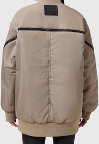 Beige Bomberjacke mit glatter Textur, gerippten Bündchen und einem Reißverschluss-Akzent über dem Rücken. Verfügt über einen schwarzen Logo-Patch am Kragen.