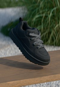Black sneaker with textured mesh upper and smooth accents. Features gray laces and rubber sole with traction pattern. Floating above wooden surface.