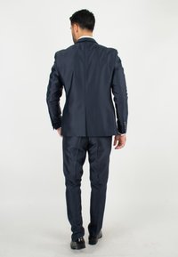 Costume bleu marine avec une coupe ajustée, une fente arrière et une texture lisse. Pantalons assortis, couleur unie, accompagnés de chaussures noires.