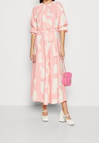 Růžové midi šaty s bílými abstraktními vzory, nařaseným výstřihem, s opaskem na zavázání a dlouhými rukávy. Slačeny s bílými botami na blokovém podpatku a malou růžovou kabelkou.