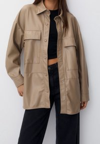 Veste-chemise en similicuir beige à coupe décontractée, avec deux poches poitrine et fermeture par boutons-pression. Portée sur un haut noir et un jean foncé.