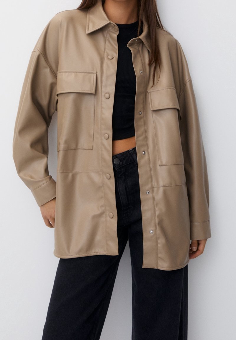 Veste-chemise en similicuir beige à coupe décontractée, avec deux poches poitrine et fermeture par boutons-pression. Portée sur un haut noir et un jean foncé.