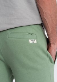 Joggers en coton vert clair avec une poche plaquée au dos, arborant une petite étiquette avec du texte. Texture douce, taille ajustée.