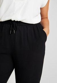 Osoba nosí volný bílý top a černé kalhoty s elastickým pasem, s rukama v kapsách, což ukazuje na neformální outfit.