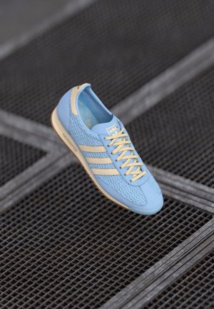 Jasnoniebieski sneaker Adidas z beżowymi paskami i sznurowadłami, unoszący się nad teksturowaną ciemną metalową siatką.