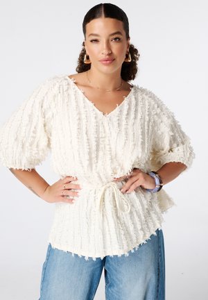 Vrouw met krullend haar draagt een getextureerde witte blouse met ceintuur in de taille, blauwe spijkerbroek, gouden oorbellen en armbanden, poseert met handen in de zij.