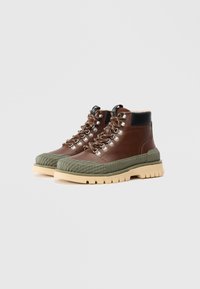 Botas altas de cuero marrón con acentos de goma verde oliva, suela texturizada estriada, ojales de metal plateado y detalle en el talón negro.