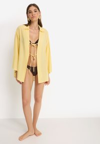 Κίτρινο ελαφρύ πουκάμισο, ανοιχτό μπροστά, με μακριά μανίκια και λεπτομέρεια ζώνης στο λαιμό, φορεμένο πάνω από ένα σκούρο καφέ μπικίνι, σε λευκό φόντο.