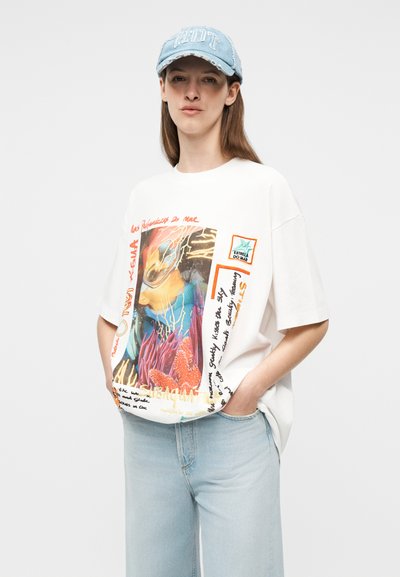 Γυναίκα που φοράει ανοιχτό μπλε καπέλο, λευκό φαρδύ μπλουζάκι με πολύχρωμο αφηρημένο σχέδιο και ανοιχτό μπλε τζιν, στέκεται με τα χέρια στις τσέπες.