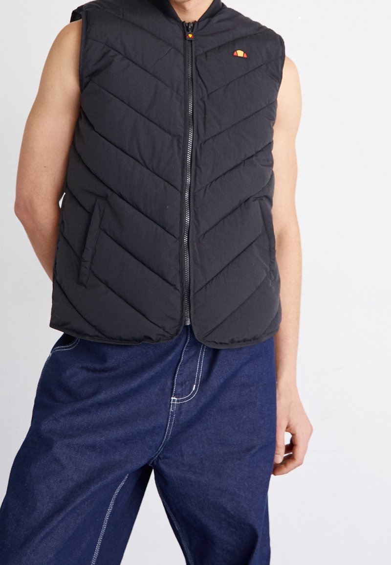 Veste sans manches noire matelassée avec fermeture éclair, portée sur un pantalon bleu foncé ample avec des coutures blanches visibles sur un fond uni.