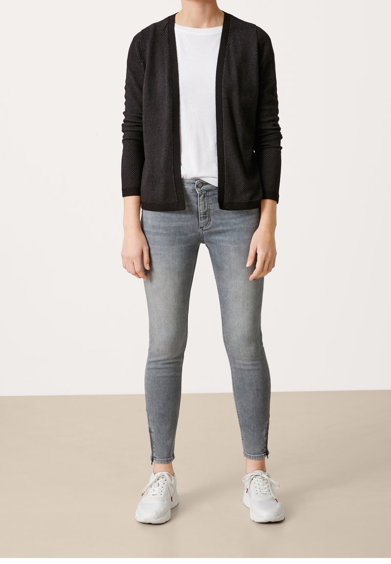 Personne portant un cardigan noir ouvert sur le devant, un t-shirt blanc, un jean skinny gris clair avec des fermetures éclair aux chevilles, et des baskets blanches se tenant sur un sol beige.