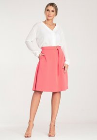 Biała bluzka z dekoltem w serek i guzikami, w połączeniu z różową spódnicą do kolan, która ma tasiemkę i okrągły pasek. Gładkie tekstury, lekki materiał.