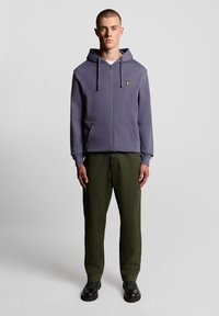 Grauer Kapuzenpullover mit durchgehendem Reißverschluss, Kordelzugkapuze, vorderen Taschen und einem kleinen gelben Logo, kombiniert mit olivgrünen Hosen und schwarzen Schuhen.