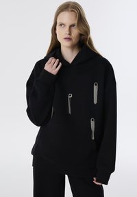 Sudadera negra con un gran bolsillo frontal, detalles en hardware plateado y borlas. Hecha de un material suave con un corte relajado.