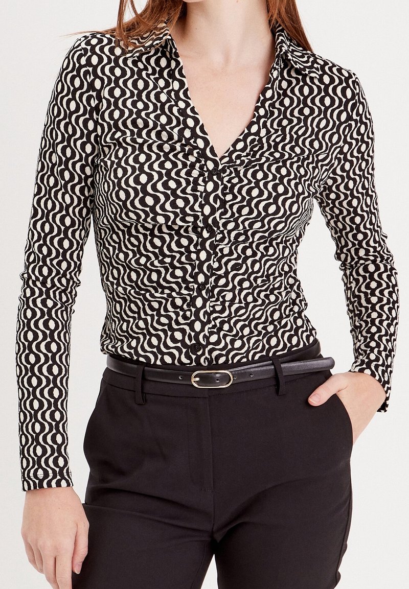 Chemise à manches longues noire et blanche avec un motif ondulé, un col à revers et des boutons. Associée à un pantalon noir ajusté et une ceinture.