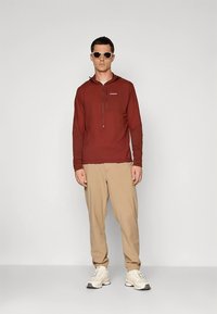 Tamsiai raudonas ilgomis rankovėmis džemperis su pusiau užtraukiamu užtrauktuku priekyje, derinamas su smėlio spalvos siaurėjančiomis kelnėmis. Modelis vilki baltus sportbačius ir saulės akinius.