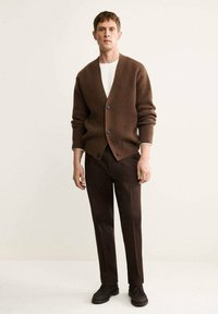 Cardigan marrone a coste con bottoni scuri, indossato sopra una camicia bianca. Abbinato a pantaloni marroni scuri e scarpe scure, in piedi contro uno sfondo chiaro.