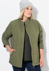 Olijfgroen gewatteerde jas met een hoge kraag, ritsluiting, twee zijzakken en een gladde textuur, gecombineerd met een grijze gebreide beanie.