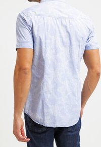 Lyhythihainen vaaleansiniinen paita, jossa on hillitty kukkakuosi, kevyestä kankaasta valmistettu, pyöristetty helma ja napituskaulus.