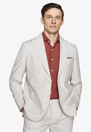 Uomo che indossa un completo beige chiaro con una camicia a bottoni color ruggine, mani nelle tasche, guarda dritto con un'espressione neutra.