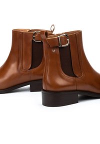 Botas de tobillo de cuero marrón con un acabado liso, paneles laterales elásticos, un detalle de hebilla y un tacón cuadrado. Costuras simples y logo de la marca visible.