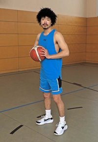 En person med en blå ermeløs skjorte og matchende shorts holder en basketball, stående på et gymgulv med lys tregulv.