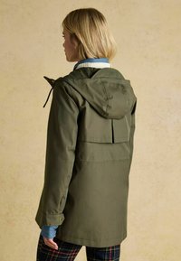 Oliwkowa kurtka z kapturem o wodoodpornej teksturze, z otworem wentylacyjnym z tyłu, zapięciem na guziki przy mankietach oraz warstwowym krojem na niebieskiej koszuli.