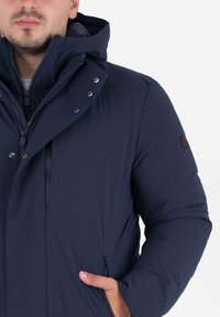 Man in een marineblauwe geïsoleerde winterjas met capuchon, dichtgeritst en geknoopt, met hand in de zijzak, tegen een effen achtergrond.
