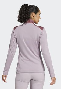 Dugim rukavima atletska majica svijetlo ljubičaste boje s rebrastom teksturom, kontrastnim tamnijim naglascima na ramenima i visokim ovratnikom. Glatka završna obrada materijala.