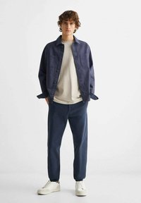 Marinblå knapp-up skjorta, ljus creamfärgad crew-neck tröja, mörka byxor och vita sneakers. Släta texturer, avslappnad passform och minimalistisk design.