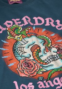 Blauwe katoenen t-shirt met een schedelontwerp, versierd met kleurrijke slangen- en rozenmotieven, ingelegd met rhinestones, en de tekst "Superdry Los Angeles" in het roze.