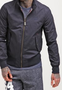 Námořnicky modrá bomber bunda se zipem vpředu a dvěma předními kapsami, s žebrovanými manžetami a límcem, vyrobená z hladkého, lehkého materiálu.