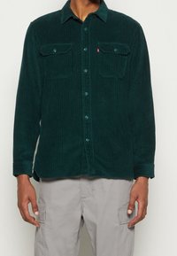 Camicia in corduroy verde scuro con bottone frontale, due tasche sul petto e maniche lunghe. Presenta una texture a coste e bottoni coordinati.