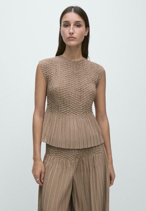 Kvinde iført ærmeløs, tekstureret brun top og matchende plisserede bukser med vidde, stående mod en ensfarvet, lys baggrund.